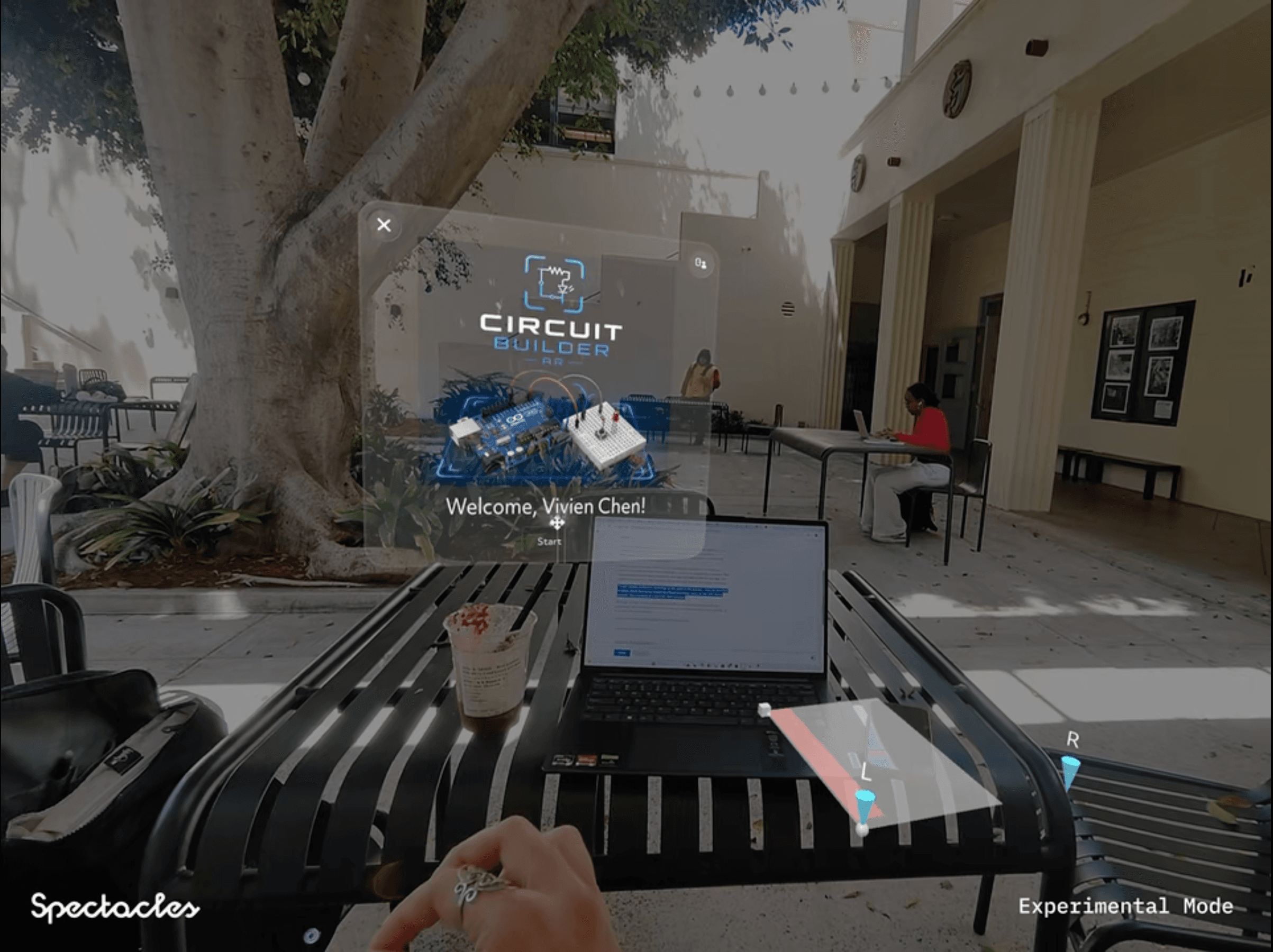 View through Snap Spectacles showing the Circuit Builder AR app welcoming a student, with a laptop visible on a table in the real world below